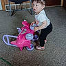 Destinee is registered to the contest to win money with this photo: toddler, child, toy, pink, car, indoor, carpet, chair, wood_paneling, door, curly_hair, white_shirt, black_pants, footwear, play, floor, room, young_child, standing, expression