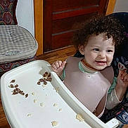 Destinee joined the competition — help win amazing prizes! baby, bib, chair, child, clothing, curly_hair, cushion, feeding, flooring, food, happy, high_chair, indoor, person, playful, room, smile, toddler, tray, wooden_door