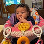 baby, infant, child, curly_hair, baby_seat, toy, finger_in_mouth, indoor, window, wooden_chair, table, blanket, clothing, polka_dots, pink, yellow, purple, face, person, cute