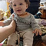 amber_necklace, baby, background, child, clothing, cozy, curly_hair, cute, face, furniture, hand, happy, home, indoors, pacifier, person, sitting, smiling, table, toy