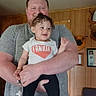 Destinee is registered to the contest to win money with this photo: child, toddler, man, holding, smiling, indoor, wood_paneling, chair, deer_head_mount, person, family, curly_hair, casual_clothing, socks, portrait, happy, living_room, home, people, baby