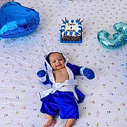 Carol is registered to the contest to win money with this photo: baby, infant, blue_clothing, boxing_outfit, bed, bedspread, patterned_sheet, balloon, cake, celebration, smile, happy, portrait, indoor, milestone, cute, child, person, lying_down, decor