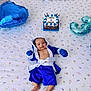 baby, infant, blue_clothing, boxing_outfit, bed, bedspread, patterned_sheet, balloon, cake, celebration, smile, happy, portrait, indoor, milestone, cute, child, person, lying_down, decor