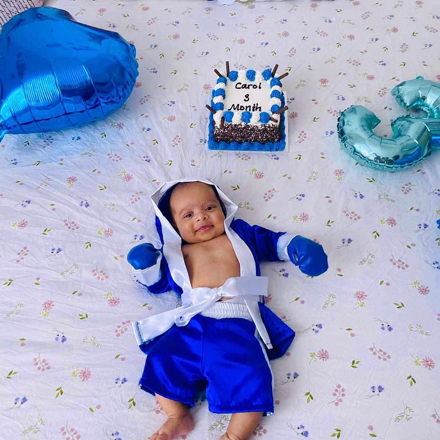 Carol is registered to the contest to win money with this photo: baby, balloon, bed, bedspread, blue_clothing, boxing_outfit, cake, celebration, child, cute, decor, happy, indoor, infant, lying_down, milestone, patterned_sheet, person, portrait, smile