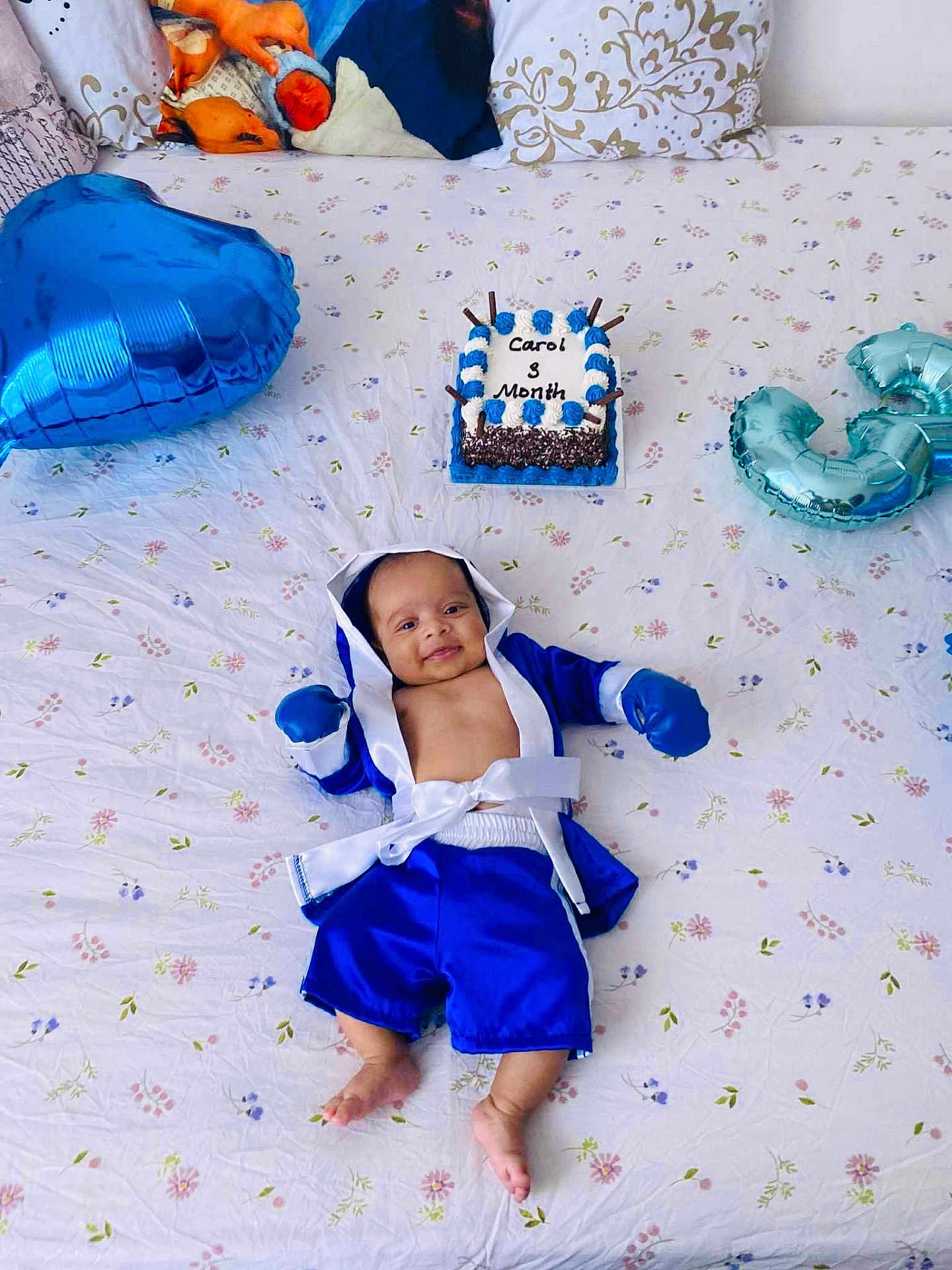 Carol is registered to the contest to win money with this photo: baby, infant, blue_clothing, boxing_outfit, bed, bedspread, patterned_sheet, balloon, cake, celebration, smile, happy, portrait, indoor, milestone, cute, child, person, lying_down, decor