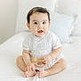 baby, child, infant, white_bed, wooden_toy, curious, cute, sitting, indoors, minimalistic, pillow, soft_lighting, portrait, clothing, person, smile, hands, feet, young, happy