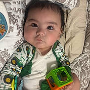 Ryan is registered to the contest to win money with this photo: baby, child, infant, pajamas, toy, bedspread, patterned, face, hand, eyes, cute, indoors, lying_down, colorful, clothing, holding, expression, young, person, play