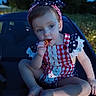 child, toddler, car, outdoor, evening, snack, food, red_hair, headband, dress, patterned_clothing, patriotic, nature, grass, trees, sitting, barefoot, face, person, portrait