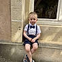 bow_tie, boy, casual, child, cobblestone, crossed_legs, daylight, fashion, ledges, outdoor, person, portrait, sandals, shirt, shorts, smile, stone_wall, suspenders, window, young