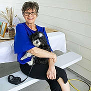 Mickey is registered to the contest to win money with this photo: black_pants, blue_top, casual, companion, concrete_floor, dog, dried_plants, glasses, happy, leash, outdoor, pet, portrait, seated, smile, table, vase, white_bench, woman, yellow_hose