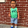 child, toddler, boy, smiling, green_tank_top, denim_shorts, blue_crocs, white_socks, brick_wall, outdoor, concrete_floor, standing, casual_clothing, short_hair, happy, cute, portrait, daylight, person, young_child