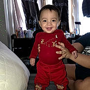 Leonardo is registered to the contest to win money with this photo: baby, child, smile, red_clothing, reindeer_design, yellow_boots, indoor, person, hand_support, curtains, clothing_covers, table, bottle, window, couch, furniture, happy, face, head, sitting