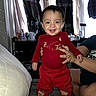 baby, child, smile, red_clothing, reindeer_design, yellow_boots, indoor, person, hand_support, curtains, clothing_covers, table, bottle, window, couch, furniture, happy, face, head, sitting