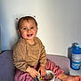 toddler, child, smiling, sitting, hair_accessory, bowl, hair_clips, pink_pants, brown_sweater, barefoot, indoors, light_wall, table, hand, face, cute, happy, playful, skin, person
