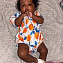 Adore is registered to the contest to win money with this photo: baby, infant, child, blanket, romper, orange_pattern, blue_leaves, sandals, bow, curly_hair, lying_down, cute, indoors, soft_texture, skin, face, hand, footwear, fashion, portrait
