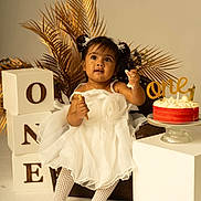 Avleen a rejoint le concours — aidez-le/la à gagner de superbes lots ! toddler, child, white_dress, birthday_cake, cake, snack, decorative_blocks, one, chair, golden_palm_leaves, studio_photo, celebration, party, portrait, cute, happy, indoor, sitting, food, festive