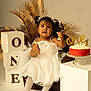 toddler, child, white_dress, birthday_cake, cake, snack, decorative_blocks, one, chair, golden_palm_leaves, studio_photo, celebration, party, portrait, cute, happy, indoor, sitting, food, festive