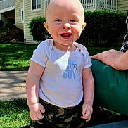 Franklin is registered to the contest to win money with this photo: baby, child, smiling, outdoor, sunlight, grass, building, hand, person, happy, clothing, camo_pants, white_shirt, face, head, portrait, standing, daylight, residential, support