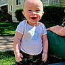 baby, child, smiling, outdoor, sunlight, grass, building, hand, person, happy, clothing, camo_pants, white_shirt, face, head, portrait, standing, daylight, residential, support