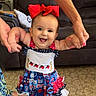 Mia joined the competition — help win amazing prizes! baby, child, infant, smiling, happy, portrait, red_bow, dress, colorful_clothing, standing, supported, hands, family, indoor, living_room, couch, laundry, carpet, fashion, photography