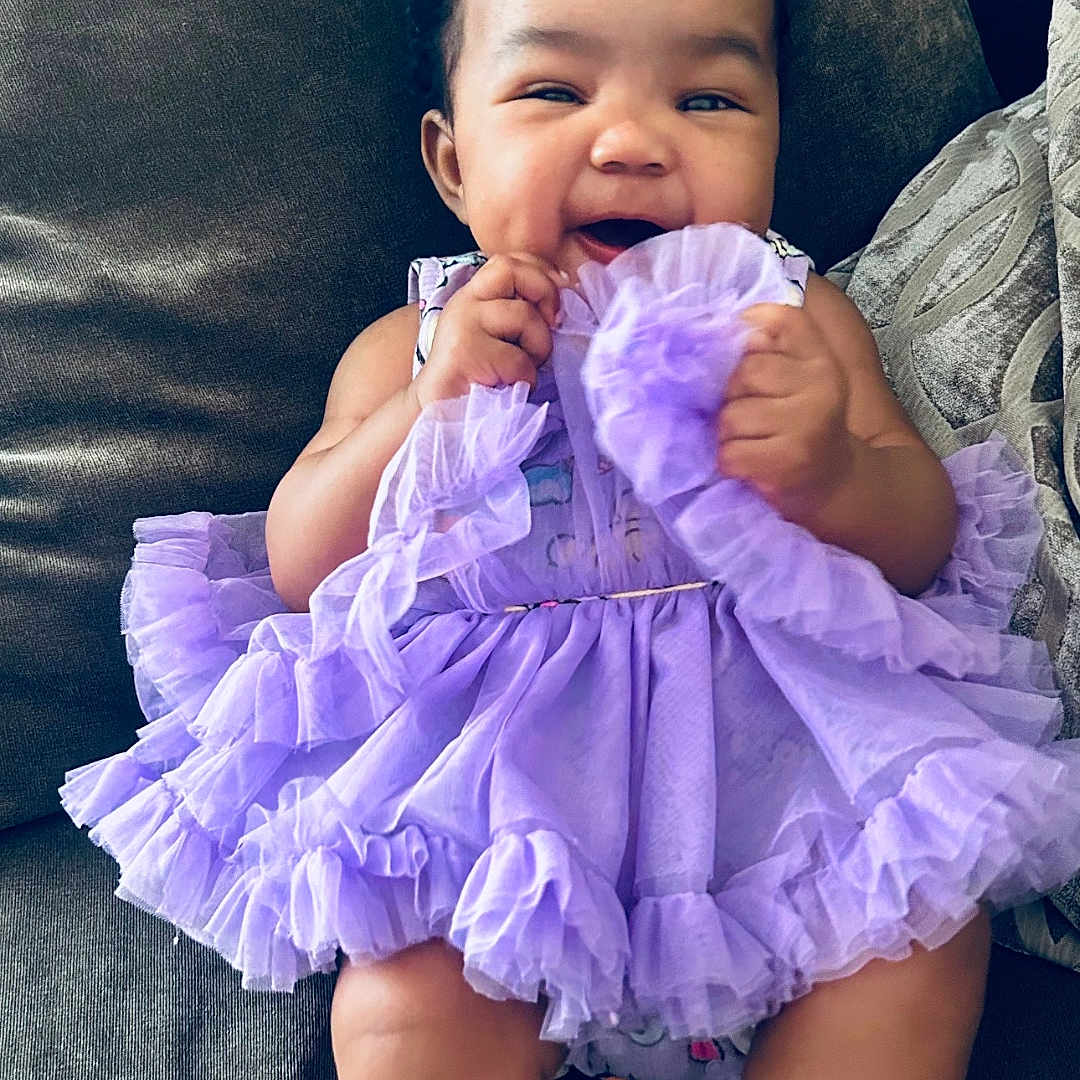 Sixx is registered to the contest to win money with this photo: baby, child, infant, smiling, happy, portrait, indoor, couch, seated, lavender_dress, ruffles, white_socks, curly_hair, face, hands, legs, baby_clothing, living_room, cute, photography