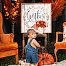 child, toddler, autumn, pumpkin, denim_overalls, standing, portrait, backward_glance, indoors, fall_decor, orange_chair, flowers, floral_arrangement, rustic_decor, framed_sign, thanksgiving, blanket, orange, home_decor, photography