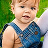 baby, child, toddler, portrait, smiling, outdoor, green_background, denim, overalls, blue_clothing, blond_hair, blue_eyes, face, close_up, sitting, being_held, adult, parent, natural_light, photography