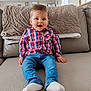 baby, child, smiling, sitting, blue_eyes, plaid_shirt, jeans, white_socks, couch, blanket, indoor, cozy, happy, portrait, person, furniture, home, casual_clothing, infant, natural_light