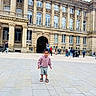 child, building, plaza, urban, people, walking, historic, architecture, outdoor, casual_clothing, hoodie, shorts, sandals, pavement, daytime, city, tourists, crowd, smiling, playful