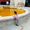 child, fountain, orange_water, hoodie, shorts, sandals, side_view, curious, urban, stone, architecture, pavement, outdoor, daylight, young_child, person, building, reflection, city, casual_clothing