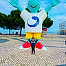 child, statue, outdoor, sky, smile, happy, sculpture, plaza, pavement, trees, person, colorful, blue_sky, art, public_space, casual_clothing, red_shoes, yellow_shorts, white_shirt, green_jacket