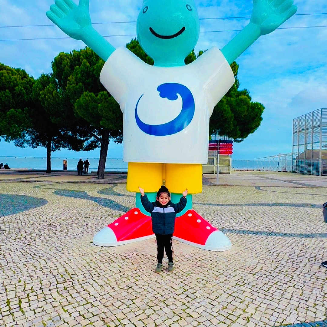 Gursher joined the competition — help win amazing prizes! child, statue, outdoor, sky, smile, happy, sculpture, plaza, pavement, trees, person, colorful, blue_sky, art, public_space, casual_clothing, red_shoes, yellow_shorts, white_shirt, green_jacket