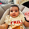 baby, child, infant, toddler, face, eyes, smile, tshirt, sun, text, hand, bracelet, seat, cushion, toy, pillow, indoor, cozy, cute, portrait