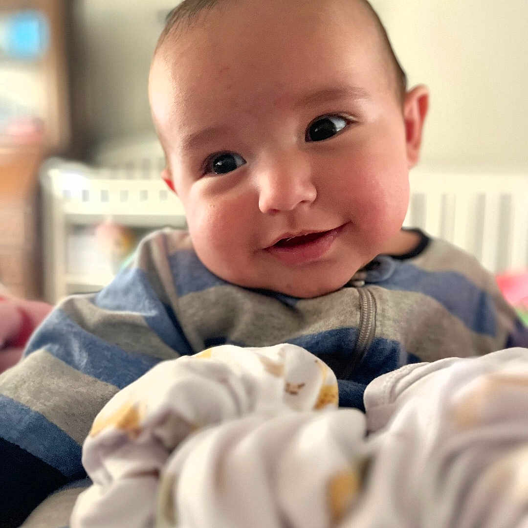 Cash is registered to the contest to win money with this photo: baby, infant, child, smiling, face, portrait, indoor, nursery, crib, blanket, blue_clothing, gray_clothing, cozy, sitting, close_up, happy, eyes, home, soft_focus, photography