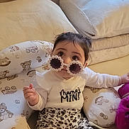 Sophia is registered to the contest to win money with this photo: baby, infant, child, sunglasses, onesie, floral_skirt, pillow, cushions, blanket, toy, stuffed_animal_pattern, window, curtain, big_eyes, dark_hair, sitting, indoor, playful, portrait, cute