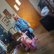 Zoè is registered to the contest to win money with this photo: child, toddler, indoor, toy, motorcycle, pink, sunglasses, denim_jacket, floor, wooden_door, table, chair, bag, slippers, home_decor, plant, picture_frame, rug, window, person