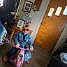 child, indoor, toy_motorcycle, red_sunglasses, denim_jacket, bag, wooden_door, table, decor, flooring, smile, footwear, curtains, wall_art, plant, light, person, play, happy, casual