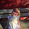 child, face_paint, bench, red_brick_wall, smile, outdoor, sunlight, wood, casual_clothing, girl, happy, portrait, daylight, colorful_shirt, green_skirt, bracelet, playful, young, cute, shadow