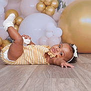 Alaiya is registered to the contest to win money with this photo: baby, infant, child, floor, wooden_floor, balloons, gold_balloons, white_balloons, headband, bow, yellow_outfit, striped_clothing, socks, celebration, party, portrait, cute, curious, indoor, photography