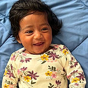 Zeel is registered to the contest to win money with this photo: baby, child, smiling, happy, infant, face, portrait, clothing, floral_pattern, onesie, blue_blanket, cute, playful, dark_hair, lying_down, indoors, skin, person, closeup, expression