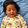 baby, child, smiling, happy, infant, face, portrait, clothing, floral_pattern, onesie, blue_blanket, cute, playful, dark_hair, lying_down, indoors, skin, person, closeup, expression