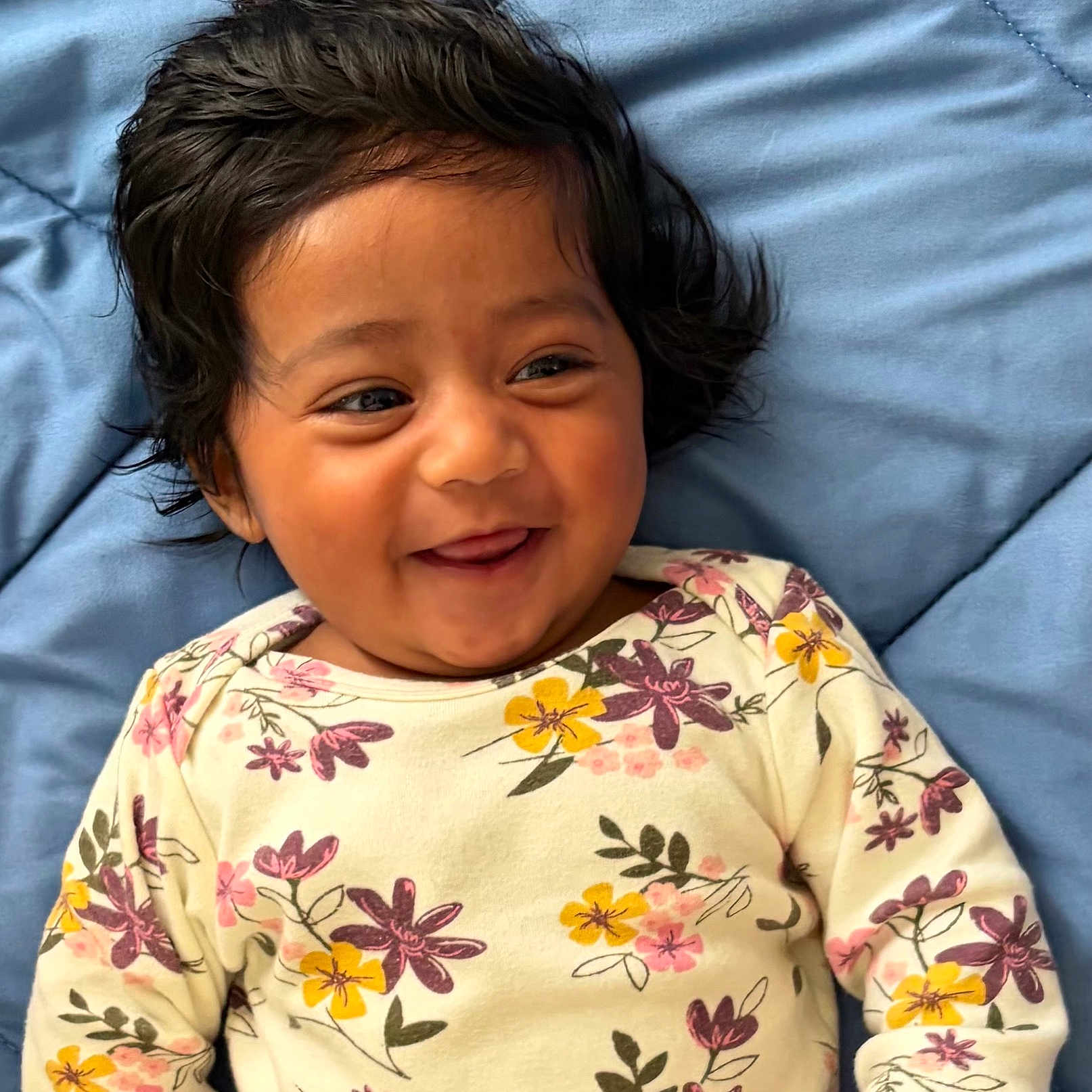 Zeel is registered to the contest to win money with this photo: baby, child, smiling, happy, infant, face, portrait, clothing, floral_pattern, onesie, blue_blanket, cute, playful, dark_hair, lying_down, indoors, skin, person, closeup, expression