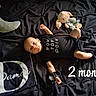 baby, infant, black_onesie, stuffed_toy, crescent_moon_pillow, blanket, bed, lying_down, milestone_text, 2_months, cute, child, soft_lighting, toy, sleeping_area, portrait, newborn, dark_background, fabric, small_child