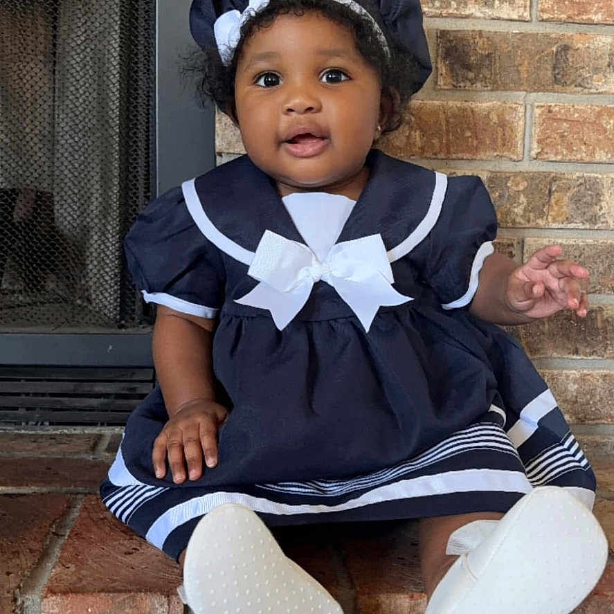Laysie is registered to the contest to win money with this photo: baby, brick_wall, child, curly_hair, cute, fireplace, footwear, happy, hat, indoors, infant, navy_blue, person, portrait, sailor_outfit, sitting, smiling, white_bow, white_shoes, young_child