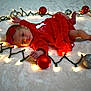baby, infant, christmas, holiday_lights, ornaments, red_dress, headband, festive, lying_down, curious, decorations, soft_surface, celebration, cute, indoors, newborn, glowing_lights, holiday_season, child, warm_lighting