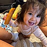 toddler, child, smiling, corn_dog, toy, blanket, indoors, happy, face, curly_hair, person, sitting, food, cozy, candid, playful, cute, baby, sock, home