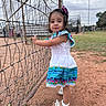 Amelia is registered to the contest to win money with this photo: child, toddler, girl, dress, bow, fence, sports_field, grass, dirt, sandals, outdoor, daylight, person, standing, looking_at_camera, curious, traditional_clothing, smile, nature, recreation