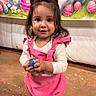 toddler, child, pink_bow, pink_overalls, easter_egg, smiling, standing, indoor, decorative_backdrop, cartoon_bunnies, colorful, festive, celebration, cute, happy, person, footwear, black_socks, white_shoes, confetti