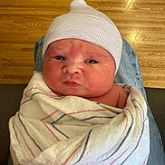 Malachi is registered to the contest to win money with this photo: newborn, baby, infant, wrapped, blanket, hat, knit_cap, person, face, head, indoors, floor, wooden_floor, seated, lap, clothing, skin, expression, portrait, cute