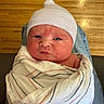 newborn, baby, infant, wrapped, blanket, hat, knit_cap, person, face, head, indoors, floor, wooden_floor, seated, lap, clothing, skin, expression, portrait, cute